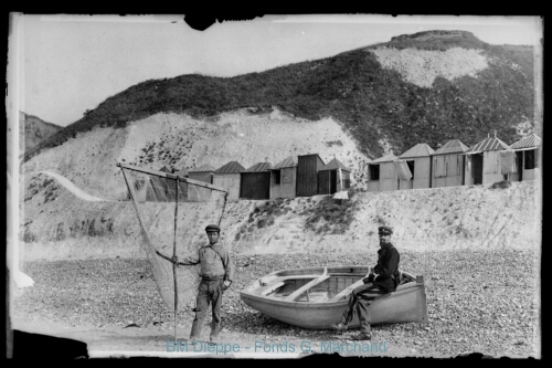 Deux pêcheurs sur les galets, avec un pousseux et une barque à sec et des cabines de plage derrière (vue de)